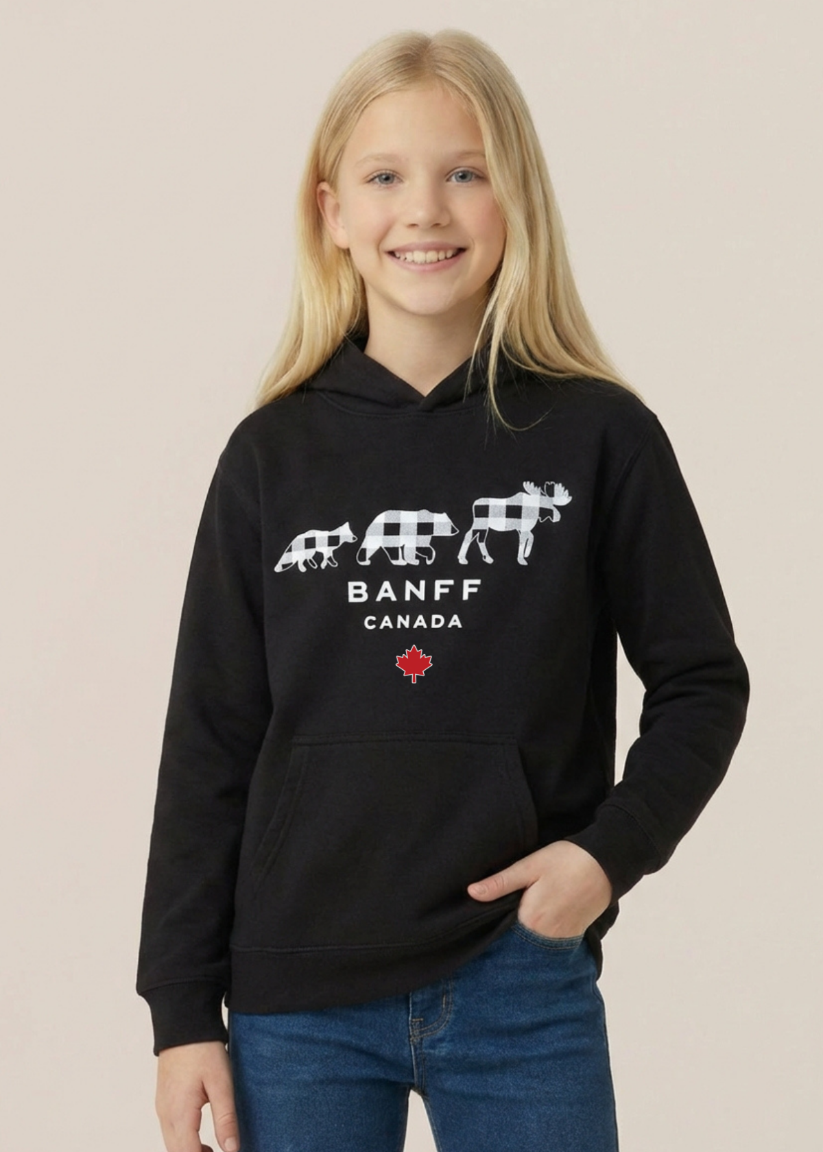 A young girl with blonde hair smiling while wearing a black pullover hoodie with a white Banff Canada graphic and blue jeans, set against a solid light beige background.