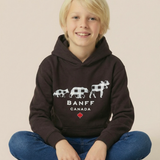 A young blonde boy sitting cross-legged wearing a dark brown Banff Canada hoodie, blue jeans, and white sneakers against a solid beige background.
