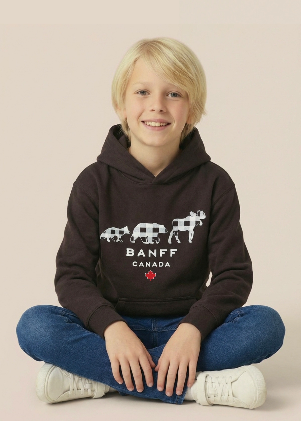 A young blonde boy sitting cross-legged wearing a dark brown Banff Canada hoodie, blue jeans, and white sneakers against a solid beige background.