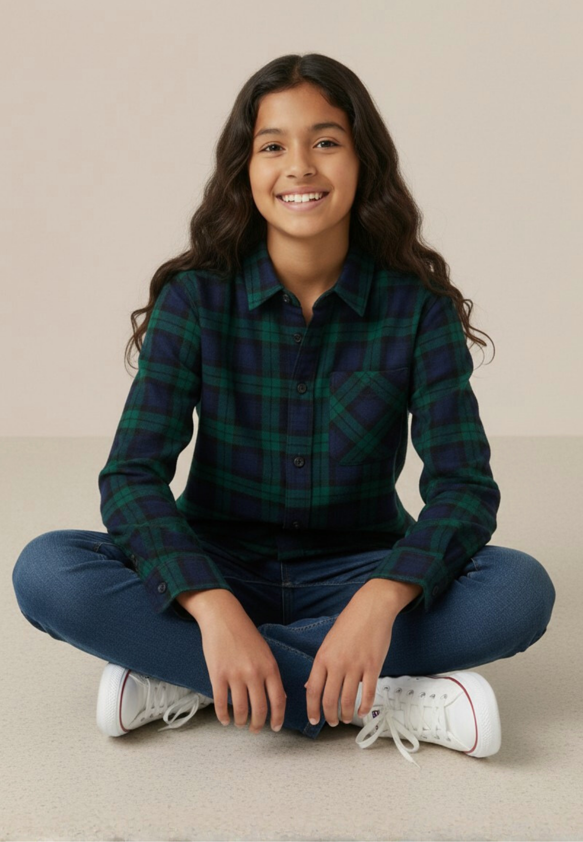 Young girl sitting cross-legged wearing a black watch tartan shirt and blue jeans on a beige background