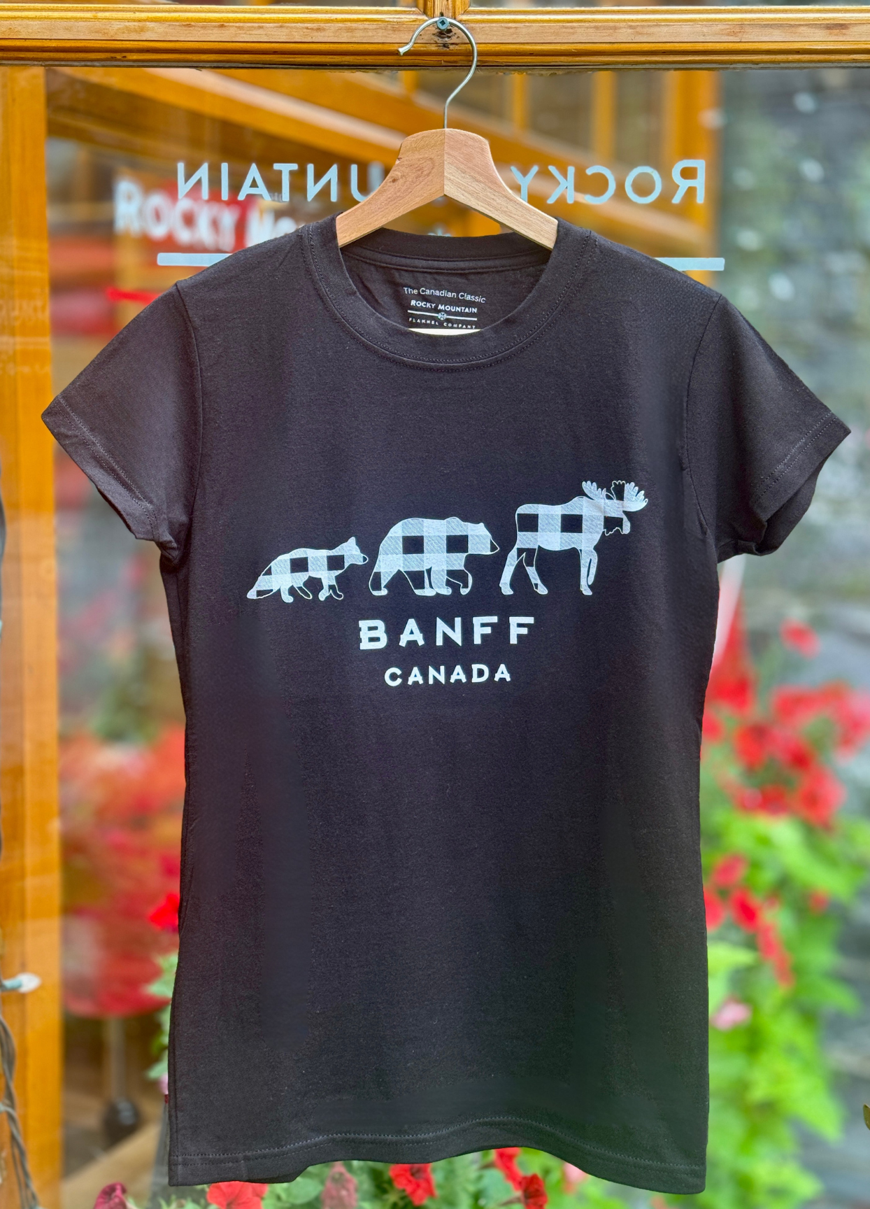 Black t-shirt with white graphic design on a hanger in front of a glass door.
