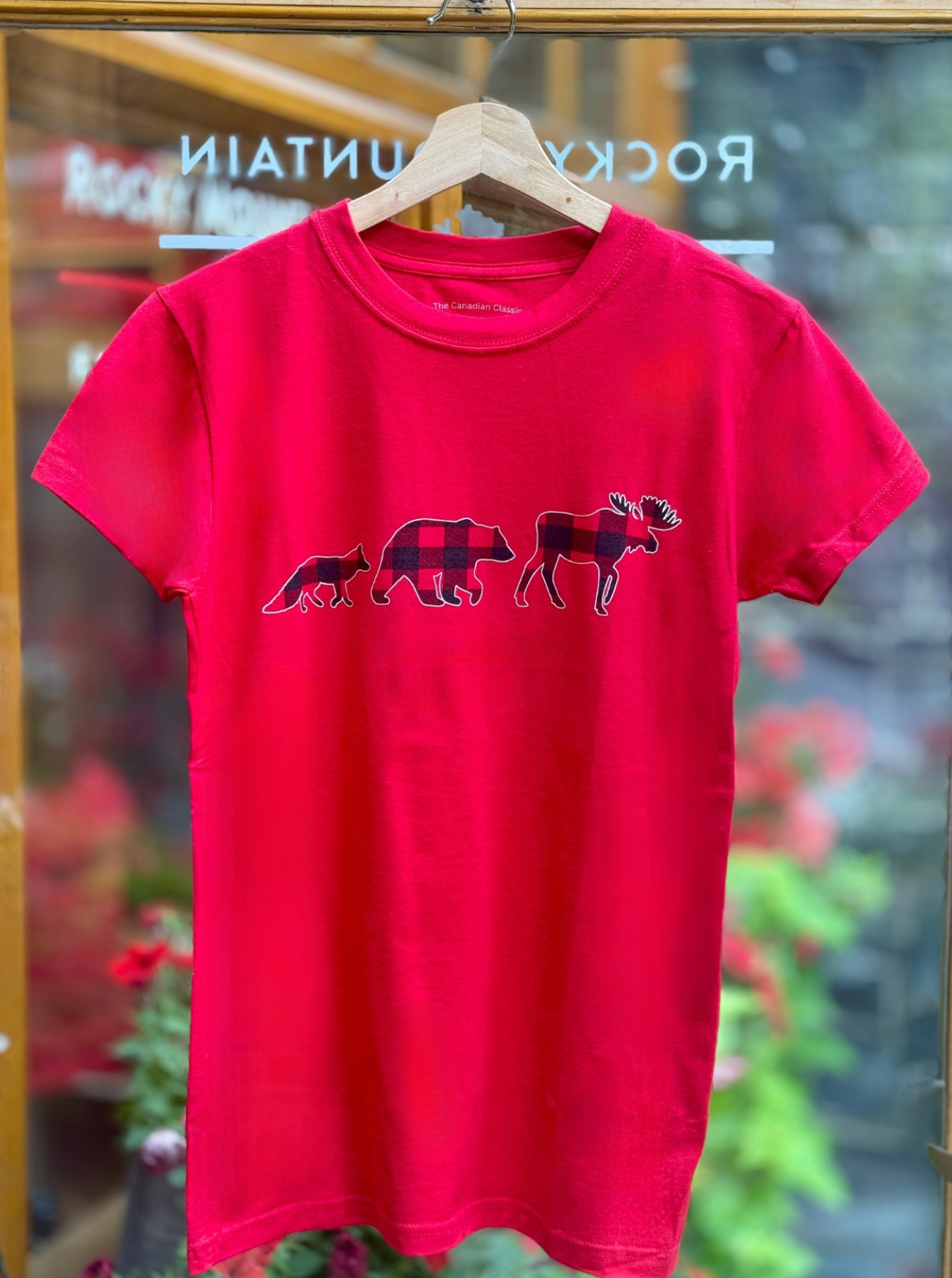 Red t-shirt with moose, bear and fox graphic on a hanger in front of a store window.