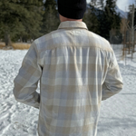 Back view of a man wearing an eggshell blue and sand plaid shirt with long sleeves, collar, and button-up front, standing outdoors.