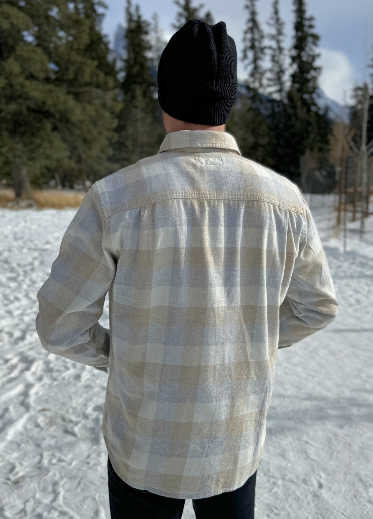 Back view of a man wearing an eggshell blue and sand plaid shirt with long sleeves, collar, and button-up front, standing outdoors.
