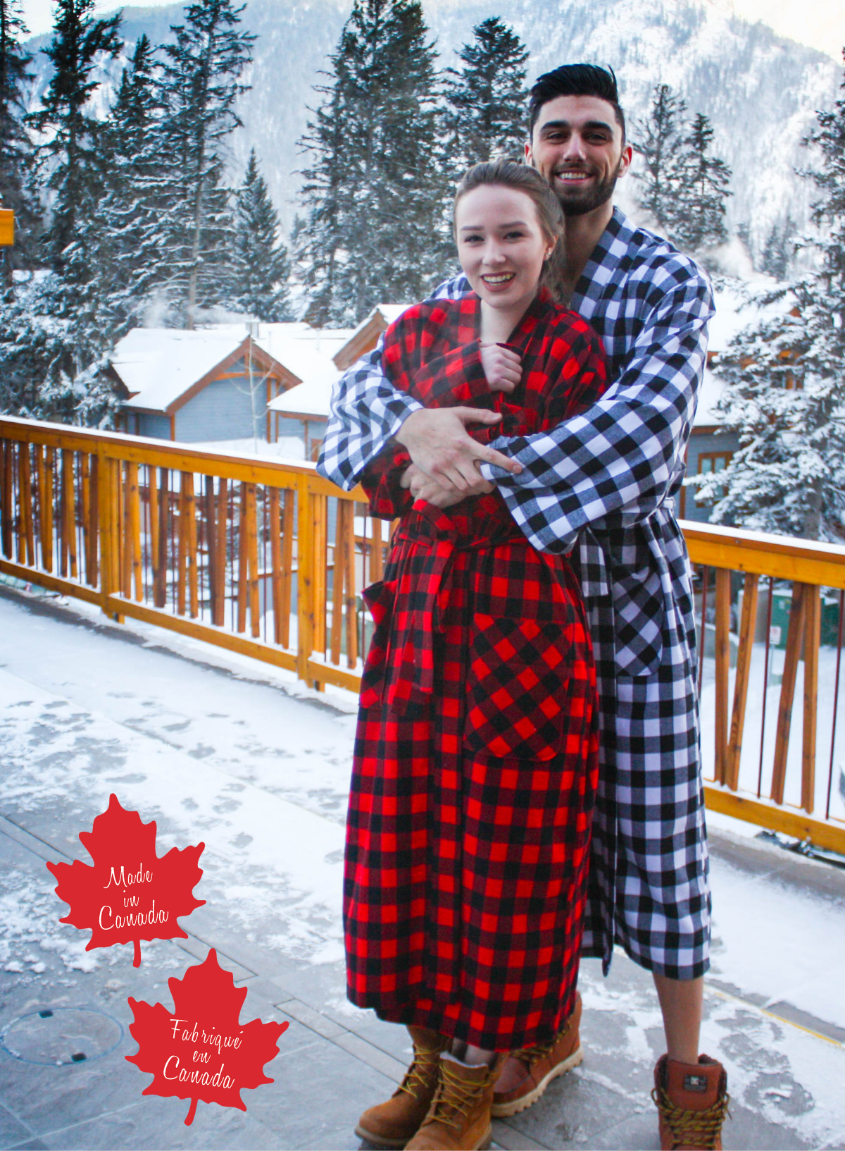 Two people in matching red and black checkered robes standing on a snowy deck with trees and mountains in the background.