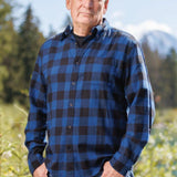 An older man with short hair wearing a long-sleeved marine blue and black buffalo check flannel shirt and blue jeans, standing outdoors.