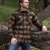 A man wearing a black and brown buffalo check flannel shirt with two flap breast pockets and two button cuff, standing next to a fallen tree.