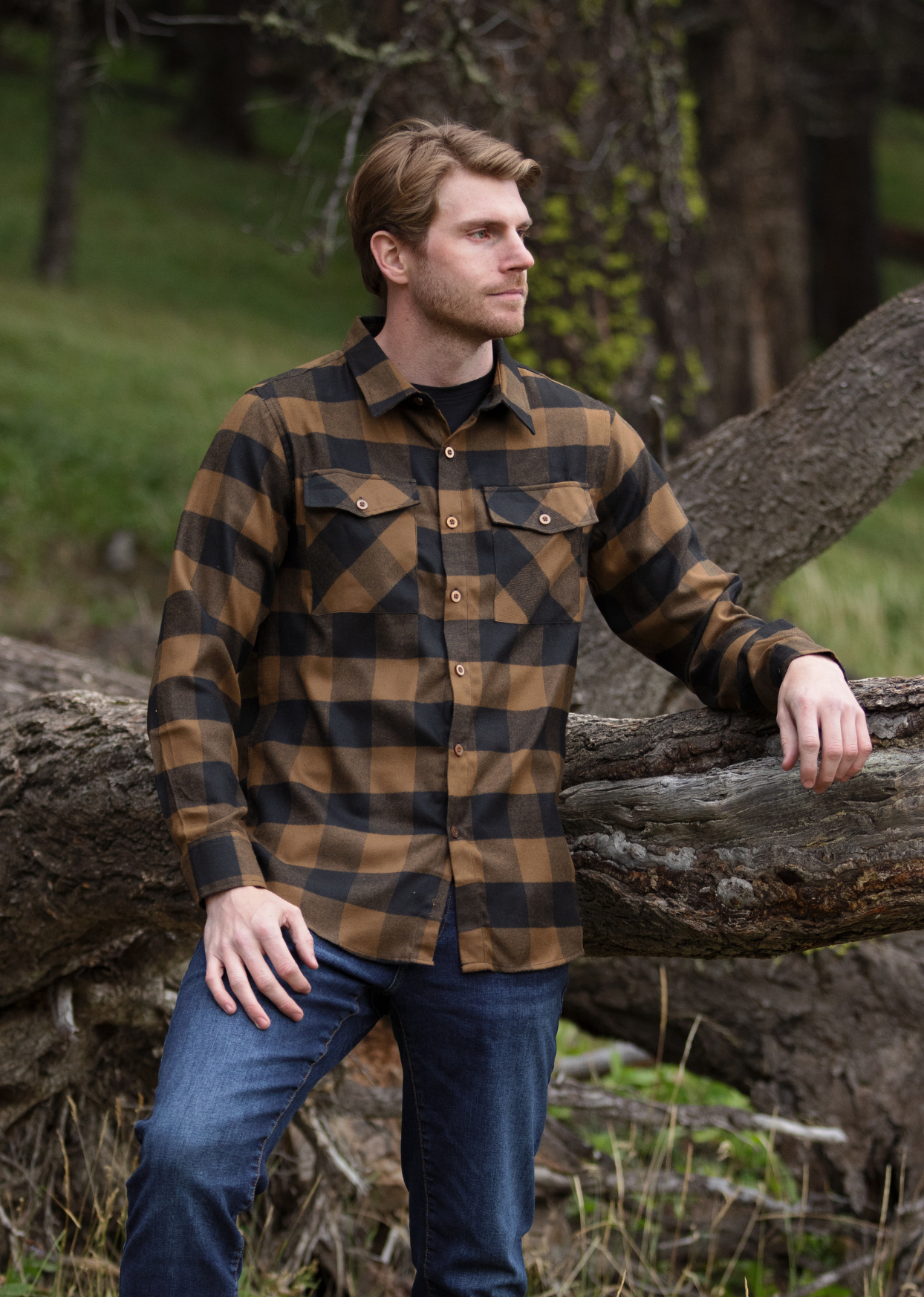 A man wearing a black and brown buffalo check flannel shirt with two flap breast pockets and two button cuff, standing next to a fallen tree.