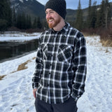 A man wearing a black and white plaid padded flannel jacket, standing near a snowy landscape.