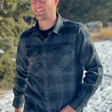 A man wearing a long-sleeved black and grey buffalo check flannel shirt with two breast pockets and a button cuff.