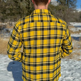 Back view of a man wearing a long-sleeved yellow and black flannel plaid shirt with two flap breast pockets and a button cuff.