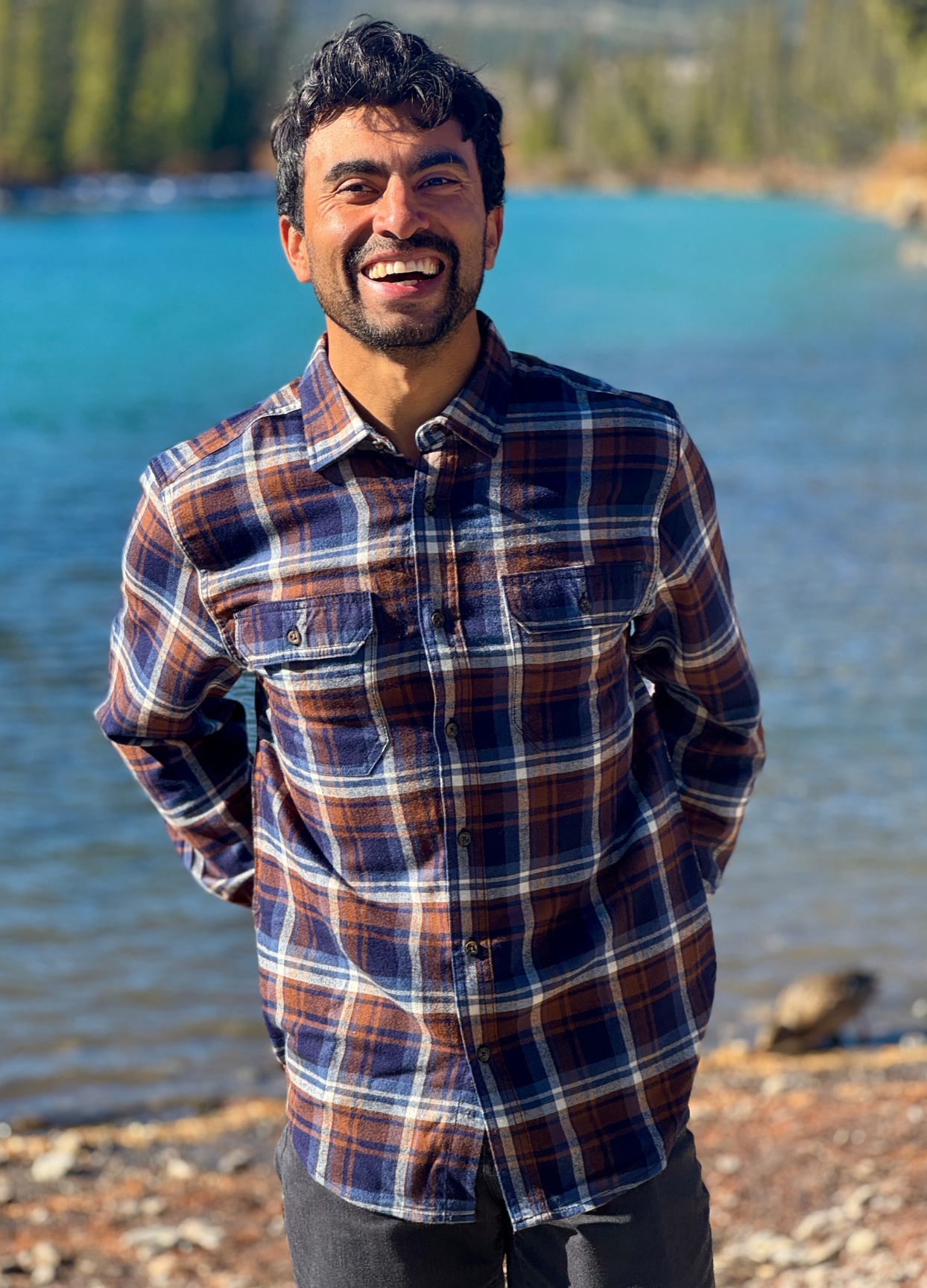 Man wearing a plaid flannel shirt standing by a river with trees in the background in Banff National Park