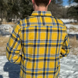 Back view of a man wearing a yellow and blue large plaid long-sleeved shirt standing outdoors.