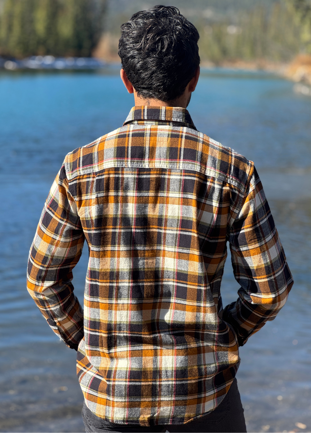 Man wearing a plaid flannel shirt by a river with trees in the background in Banff National Park