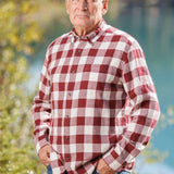 An older man with short grey hair wearing a burgundy and white buffalo check flannel shirt and blue jeans, standing near a body of water.