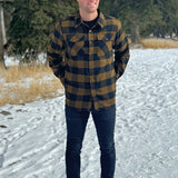 A man wearing a black and brown buffalo check flannel shirt with two flap breast pockets and two button cuff, standing with his hands in his back pockets.