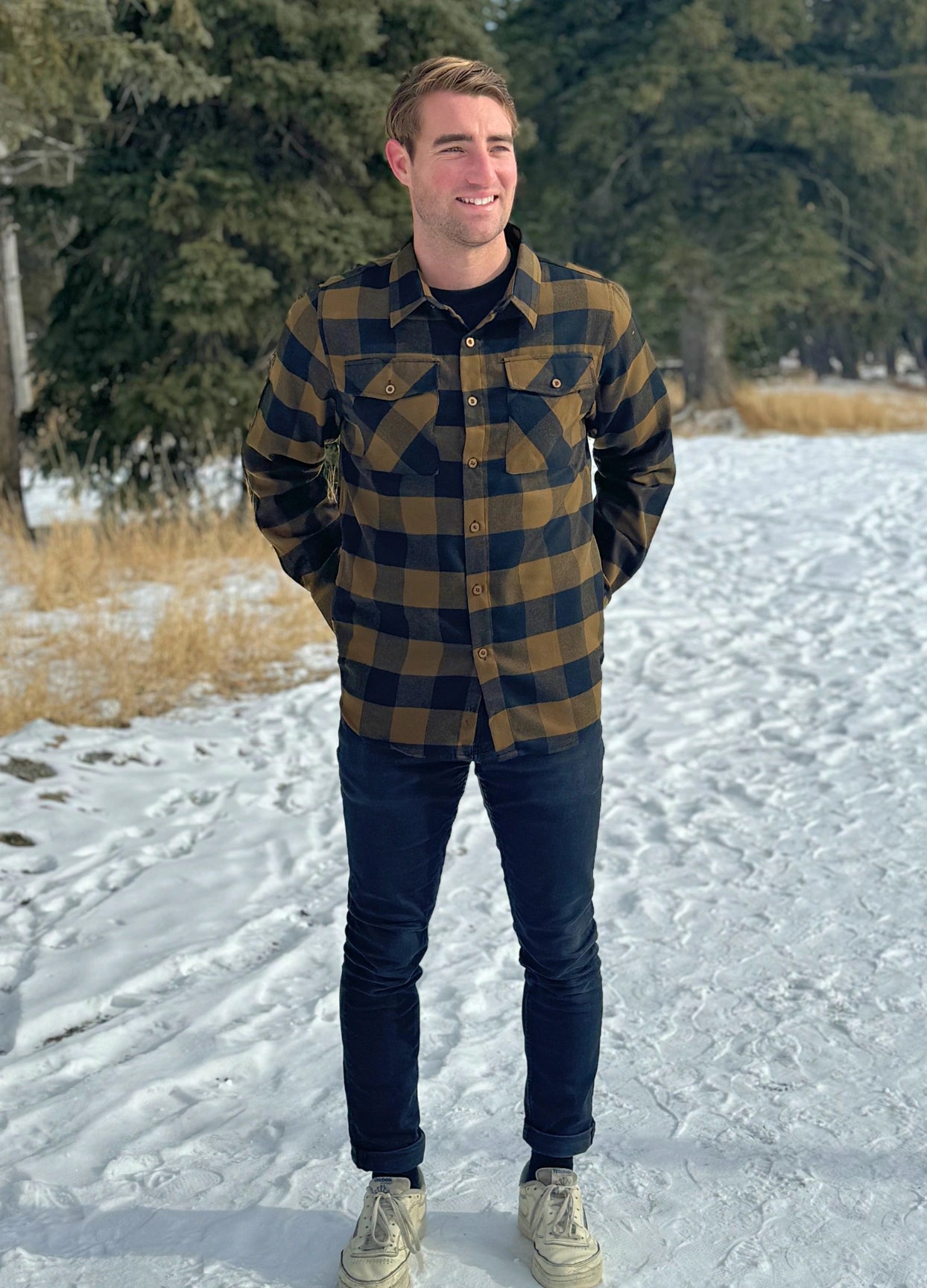 A man wearing a black and brown buffalo check flannel shirt with two flap breast pockets and two button cuff, standing with his hands in his back pockets.