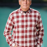 An older man with short grey hair wearing a burgundy and white buffalo check flannel shirt and blue jeans, standing near a body of water.
