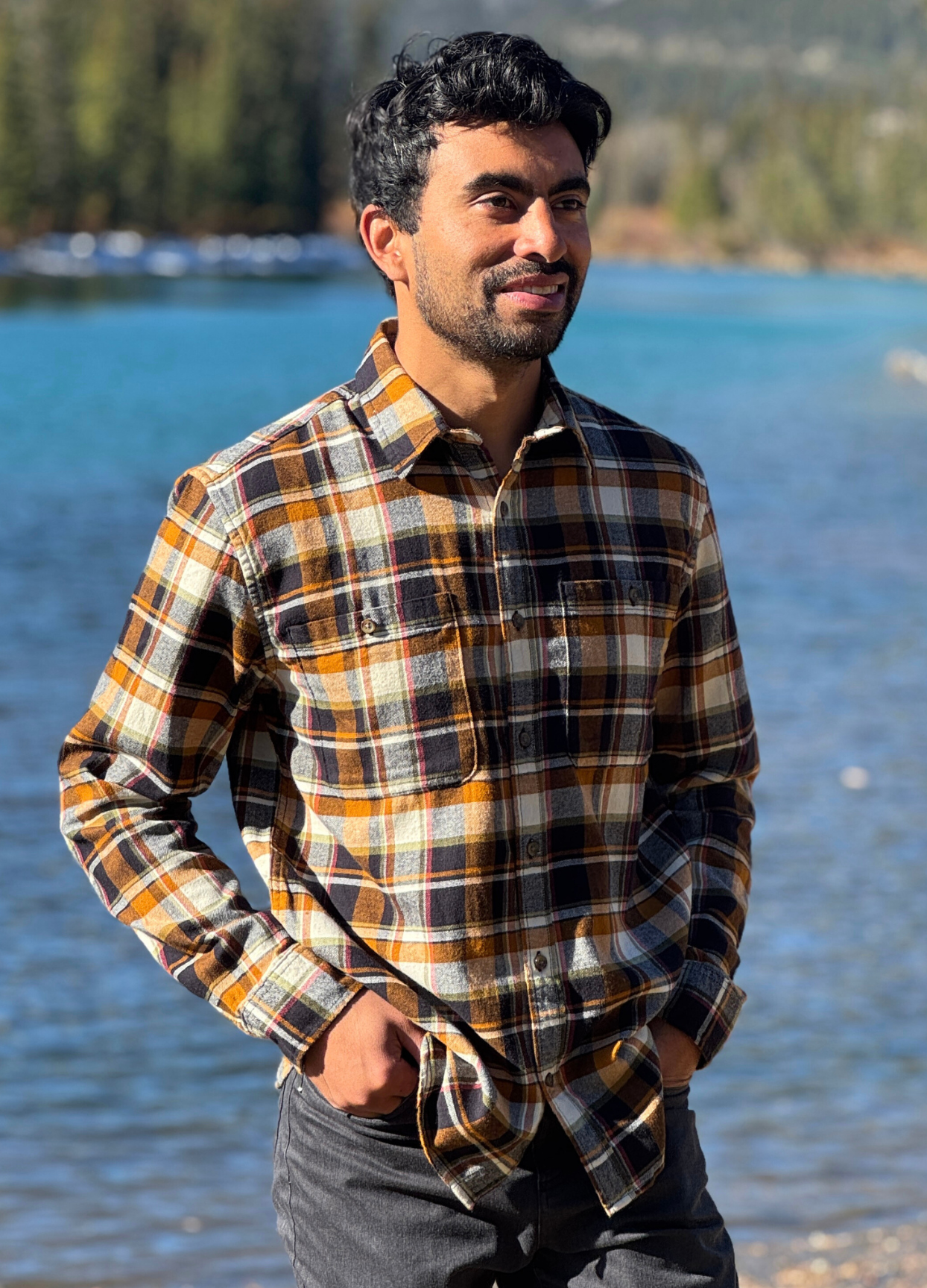 Man wearing a plaid flannel shirt standing by a river with trees in the background in Banff National Park