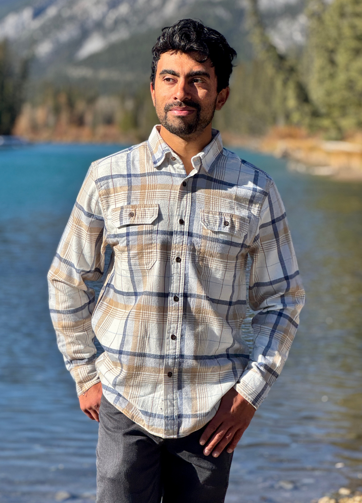 Man wearing a plaid flannel shirt standing by a river with mountains in the background in Banff National Park