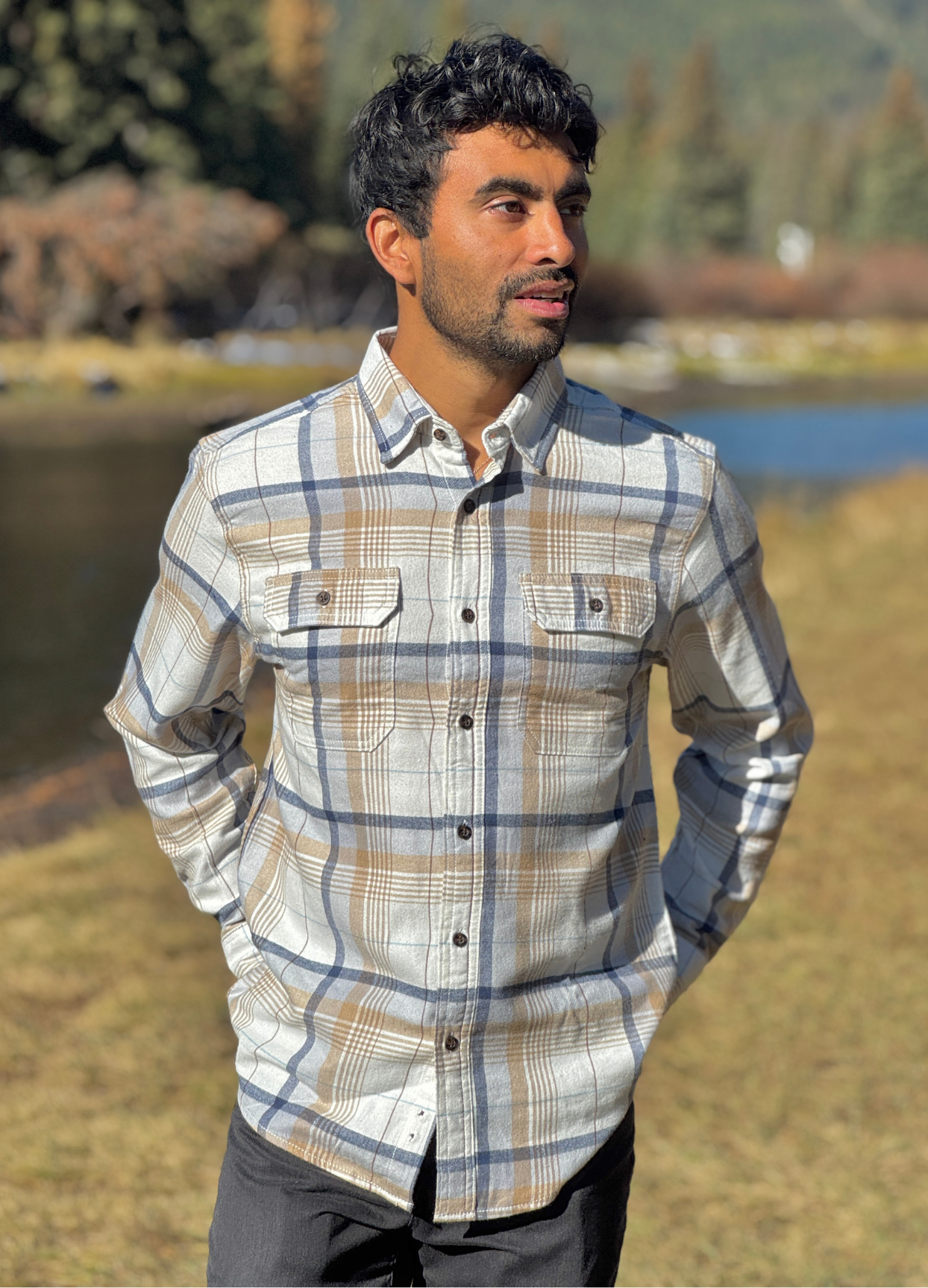 Man wearing a plaid flannel shirt standing outdoors with a river and trees in the background in Banff National Park
