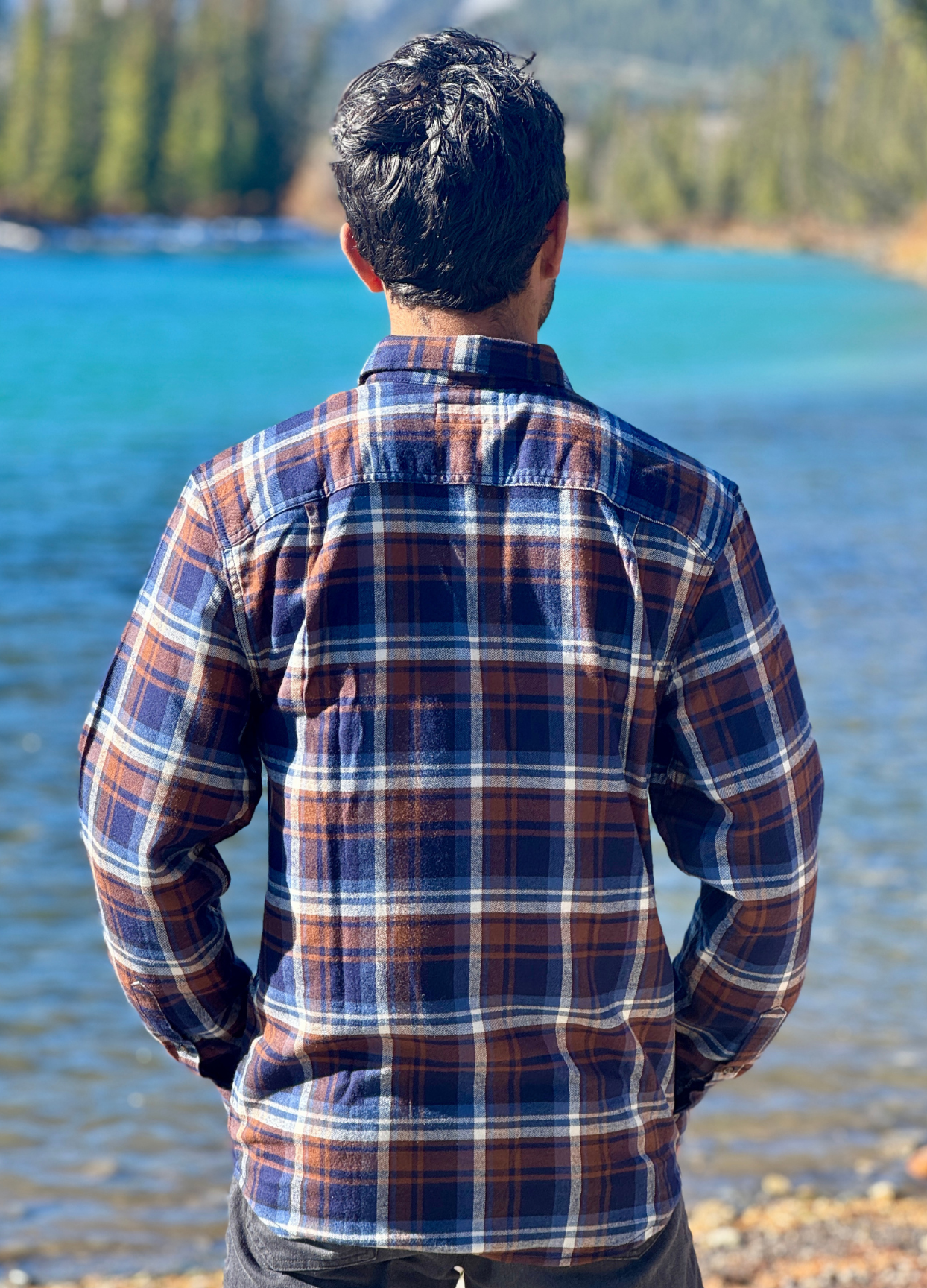 Person wearing a plaid flannel shirt standing by a river with mountains in the background in Banff National Park