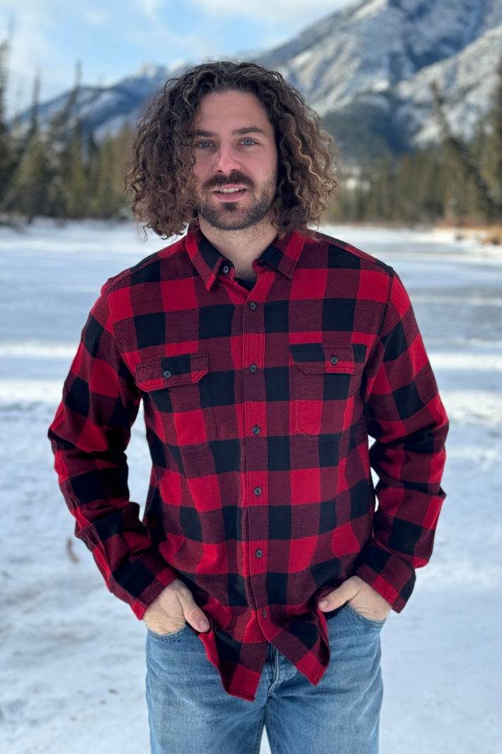 A man standing outdoors in a snowy landscape, wearing a red and black large buffalo check flannel shirt with long sleeves, button-up collar, and front pockets. This shot is taken in Banff National Park