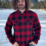 A man standing outdoors in a snowy landscape, wearing a red and black large buffalo check flannel shirt with long sleeves, button-up collar, and front pockets. This shot is taken in Banff National Park