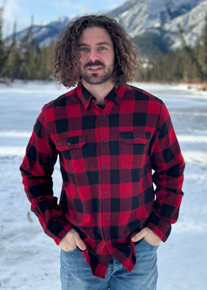 A man standing outdoors in a snowy landscape, wearing a red and black large buffalo check flannel shirt with long sleeves, button-up collar, and front pockets. This shot is taken in Banff National Park