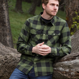 A man wearing a long-sleeved black and olive buffalo check flannel shirt with two flap breast pockets and a shirt tail hemline, standing outdoors. He is leaning up against a tree branch. He has paired the olive shirt with blue denim jeans.