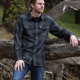 A man wearing a black and grey buffalo check flannel shirt with two flap breast pockets and two button cuff, standing next to a fallen tree in a natural setting.