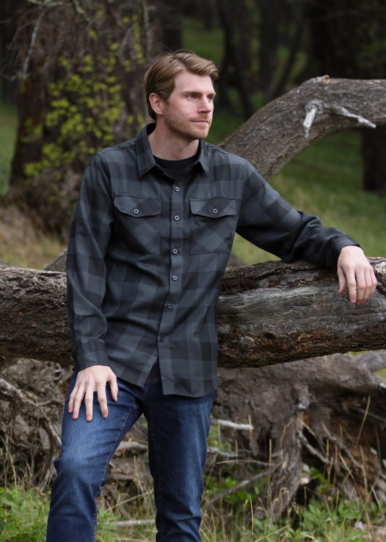 A man wearing a black and grey buffalo check flannel shirt with two flap breast pockets and two button cuff, standing next to a fallen tree in a natural setting.