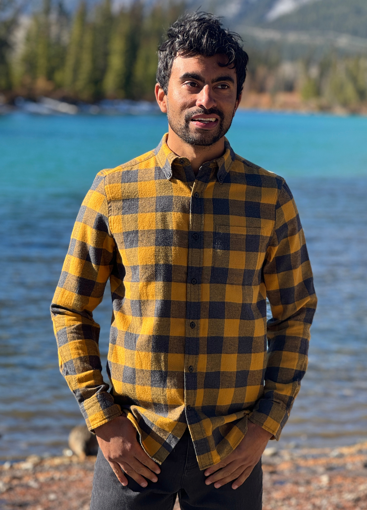 Man wearing a gold and grey plaid flannel shirt standing by a river with mountains in the background in Banff National Park