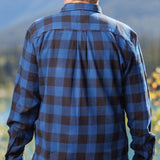 Back view of a man wearing a long-sleeved marine blue and black buffalo check flannel shirt and blue jeans, standing outdoors.