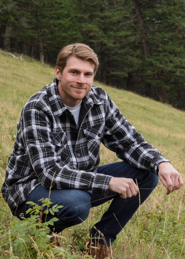 Man in a plaid jacket and jeans squatting in a grassy field with trees and mountains in the background