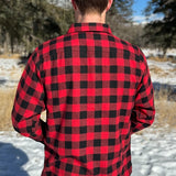 Back view of a man wearing a red and black buffalo check flannel shirt with two flap breast pockets and a button cuff, standing outdoors.