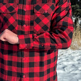 Close up of man wearing a red and black buffalo check flannel shirt with two flap breast pockets and a button cuff, standing outdoors.