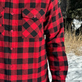 Close up of a man wearing a red and black buffalo check flannel shirt with two flap breast pockets and a button cuff, standing outdoors.