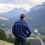 Back view of man wearing a royal blue and black buffalo check padded flannel camping outdoor jacket with jeans. There are mountains in the background