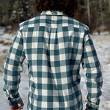 Back view of a man standing outdoors wearing a teal buffalo check flannel shirt with long sleeves, button-up collar, and a single breast pocket.