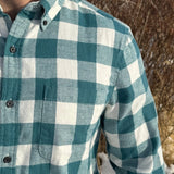 Close up of a man standing outdoors wearing a teal buffalo check flannel shirt with long sleeves, button-up collar, and a single breast pocket.