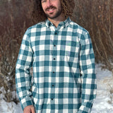 A man standing outdoors wearing a teal buffalo check flannel shirt with long sleeves, button-up collar, and a single breast pocket.