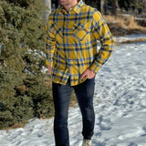 A man wearing a yellow and blue large plaid long-sleeved shirt standing outdoors.