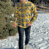 A man wearing a yellow and blue large plaid long-sleeved shirt standing outdoors.