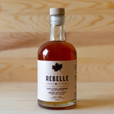 A bottle of Rebelle brand maple syrup with a wooden cap, placed on a wooden surface.