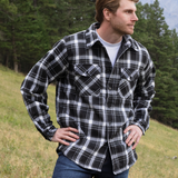 A man wearing a long-sleeved black and white plaid flannel jacket with a shirttail hemline, side pockets, and 2 button cuffs, standing outdoors.