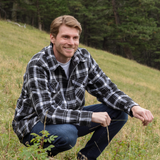 A man wearing a long-sleeved black and white plaid flannel jacket with a shirttail hemline, side pockets, and 2 button cuffs, sitting outdoors.