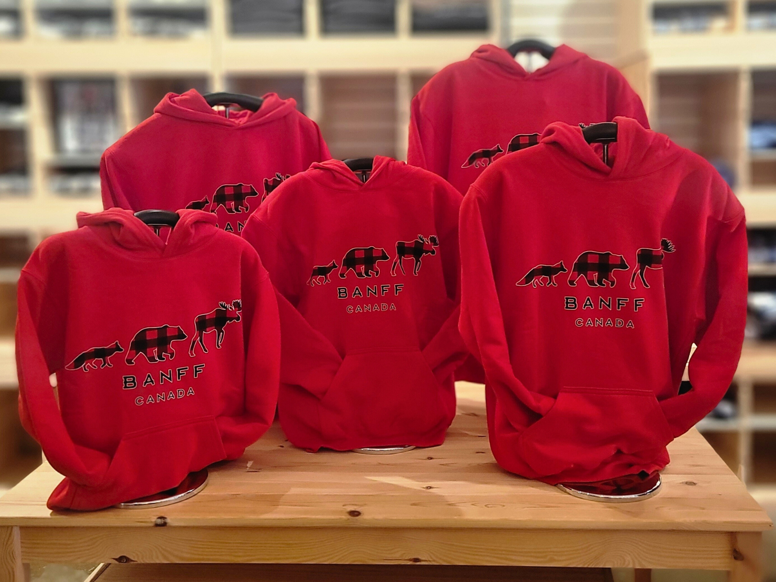 Red kids hoodies with a logo and 'Banff Canada' text on a wooden table. Showing the various kids sizes.