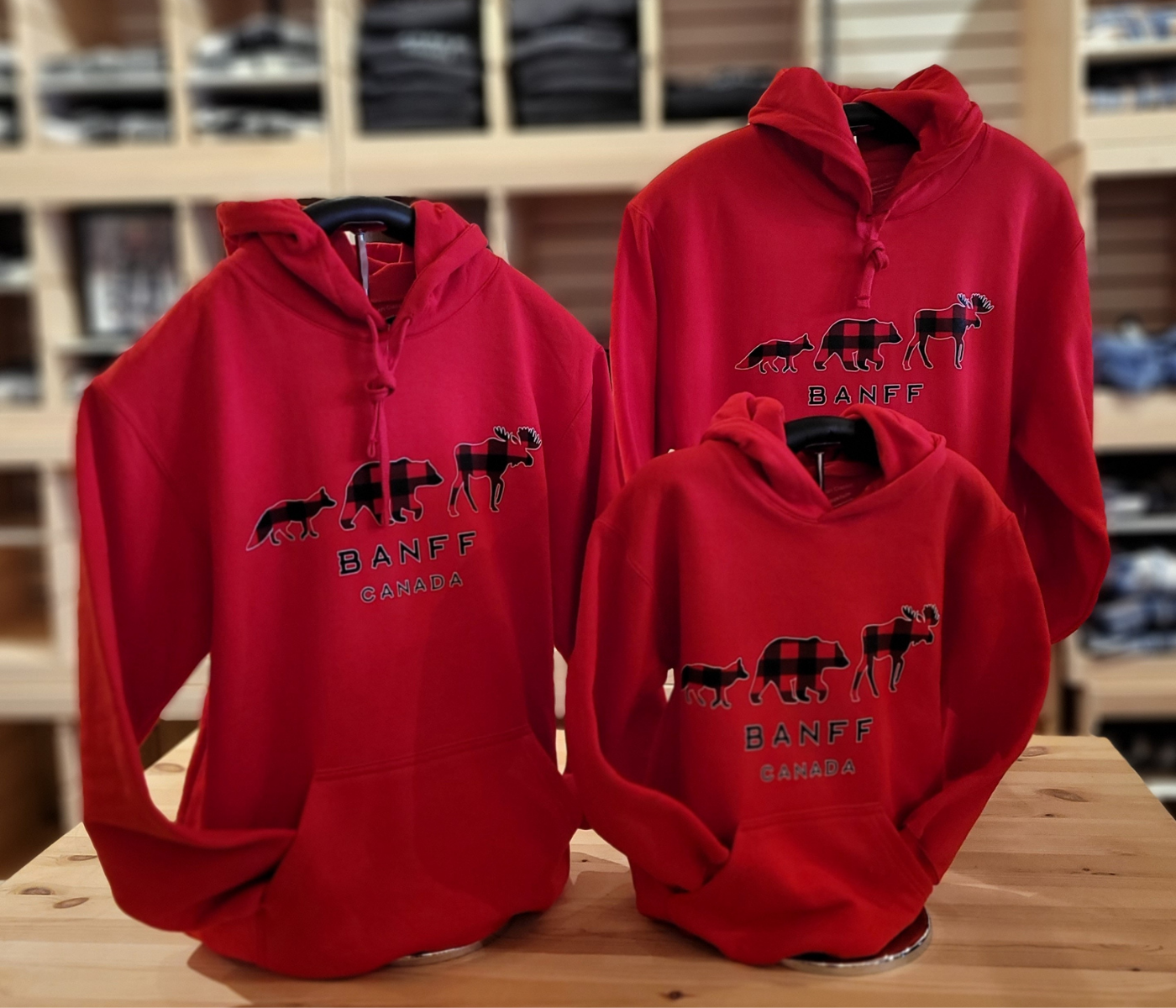 Three red hoodies with 'Banff Canada' design on a wooden surface. Featuring three iconic Canadian animals - Bear, Moose and Fox