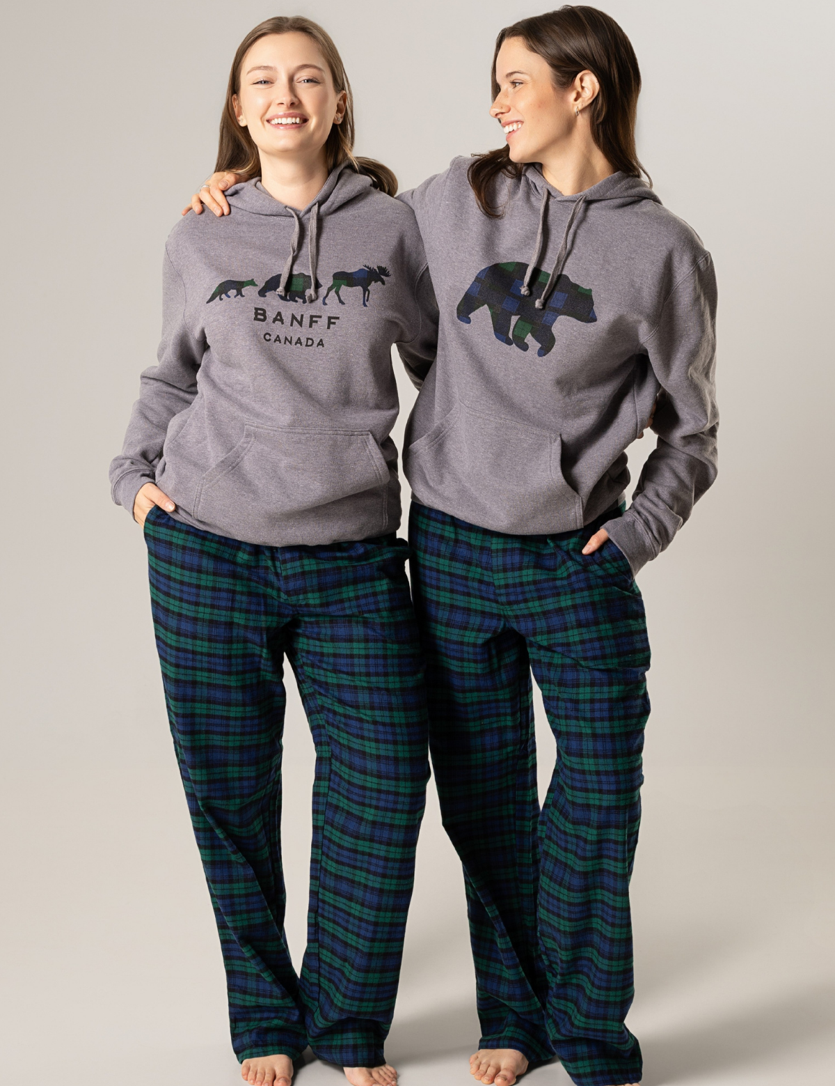 Two people wearing matching gray hoodies with a bear design and 'Banff Canada' text, paired with plaid pants on a plain background.